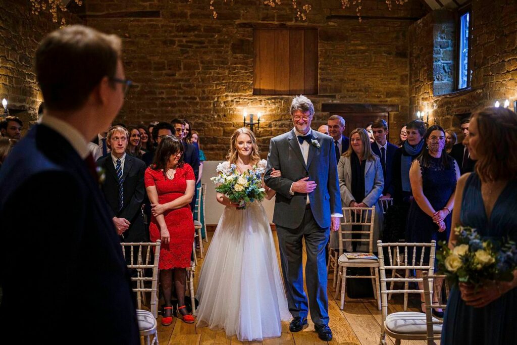 A bride in a white dress, holding a bouquet, walks down the aisle with her father in a gray suit. Guests seated on wooden chairs watch, and an officiant stands on the left. The setting is a rustic, warmly-lit venue with stone walls and wooden floors. Image by Documentary Wedding Photographer Lewis Bishop Photography.