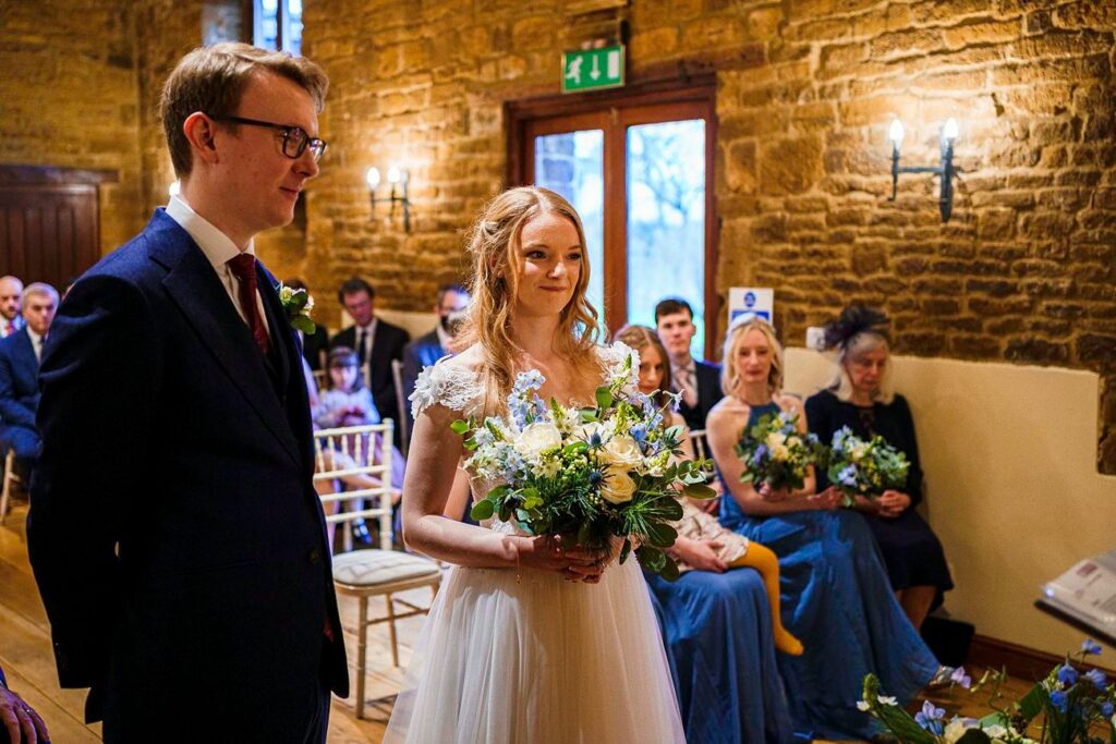 A bride and groom stand at the altar during a wedding ceremony inside a rustic venue with stone walls. The bride holds a bouquet of flowers, and wedding guests, some holding bouquets, are seated in the background, watching the couple. Image by Documentary Wedding Photographer Lewis Bishop Photography.