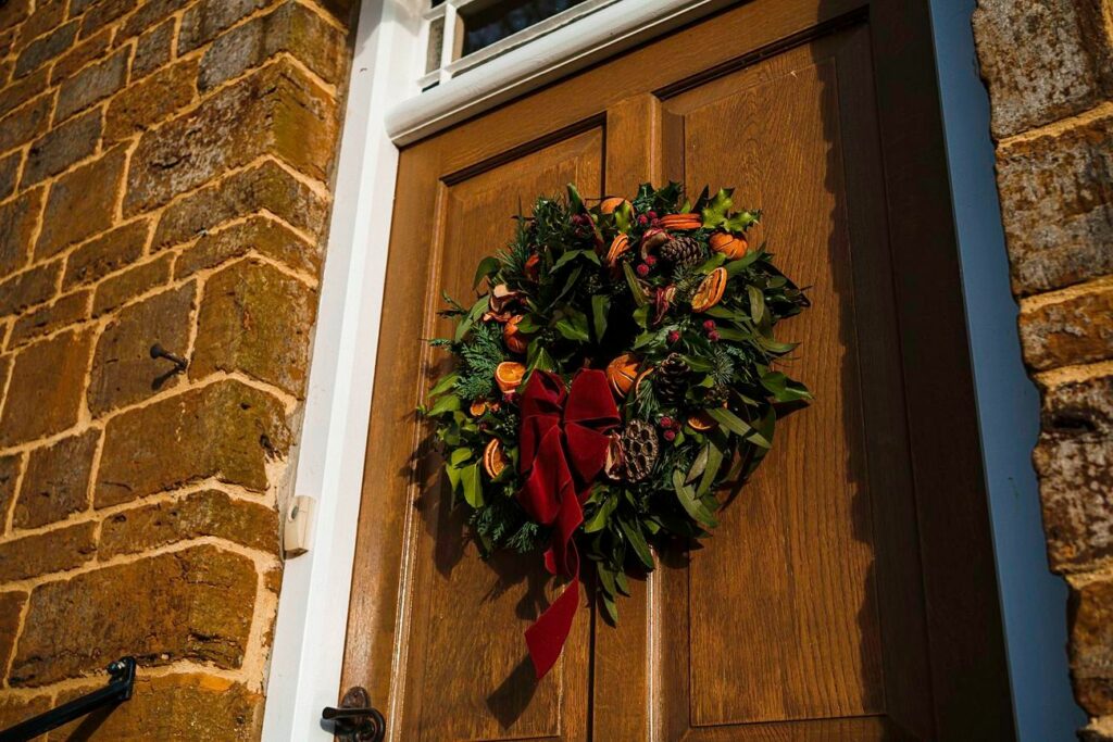 A festive wreath adorned with pinecones, berries, dried orange slices, and a large red bow hangs on a brown wooden door. The door is part of a brick building with rustic, textured walls. Image by Documentary Wedding Photographer Lewis Bishop Photography.