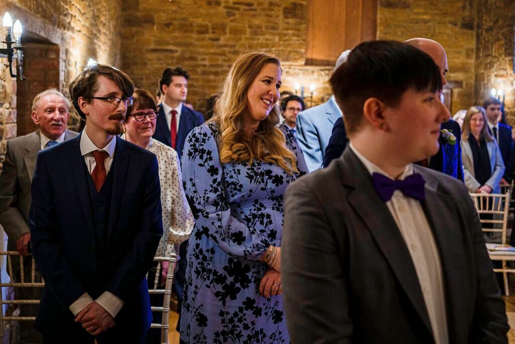 People are standing in rows inside a warmly lit venue with stone walls and wooden decor. In the foreground, three individuals stand out: a person with a suit and glasses, a smiling individual in a blue floral dress, and a person with short hair wearing a suit and purple bow tie. Image by Documentary Wedding Photographer Lewis Bishop Photography.