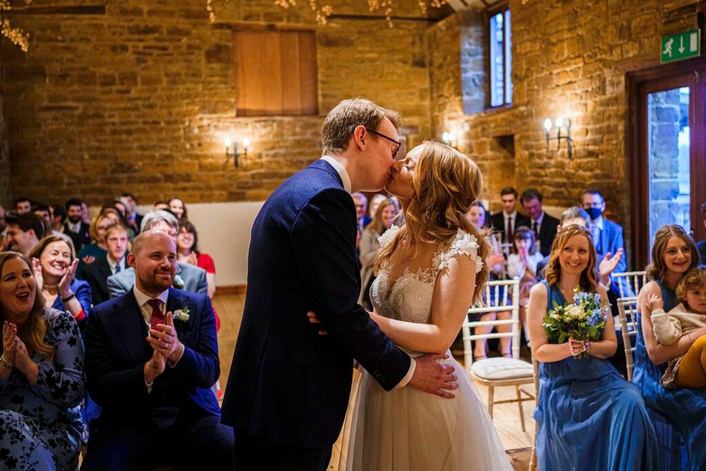 A bride and groom share a kiss at their wedding ceremony in a cozy venue with stone walls. Guests are seated on either side, clapping and smiling. The bride wears a white gown, and the groom is in a dark suit. Bridesmaids in blue dresses sit to the right. Image by Documentary Wedding Photographer Lewis Bishop Photography.