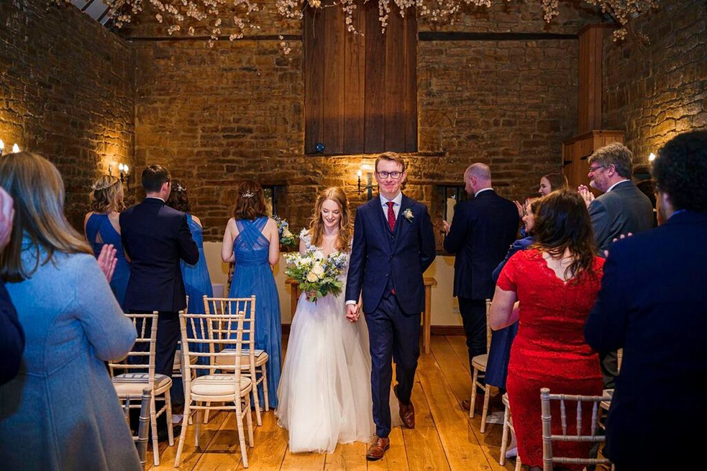 A newlywed couple holding hands and smiling, walks down the aisle in a rustic venue adorned with fairy lights and flowers. Guests in formal attire stand and clap. The bride wears a white gown and holds a bouquet; the groom is in a navy suit with a red tie. Image by Documentary Wedding Photographer Lewis Bishop Photography.