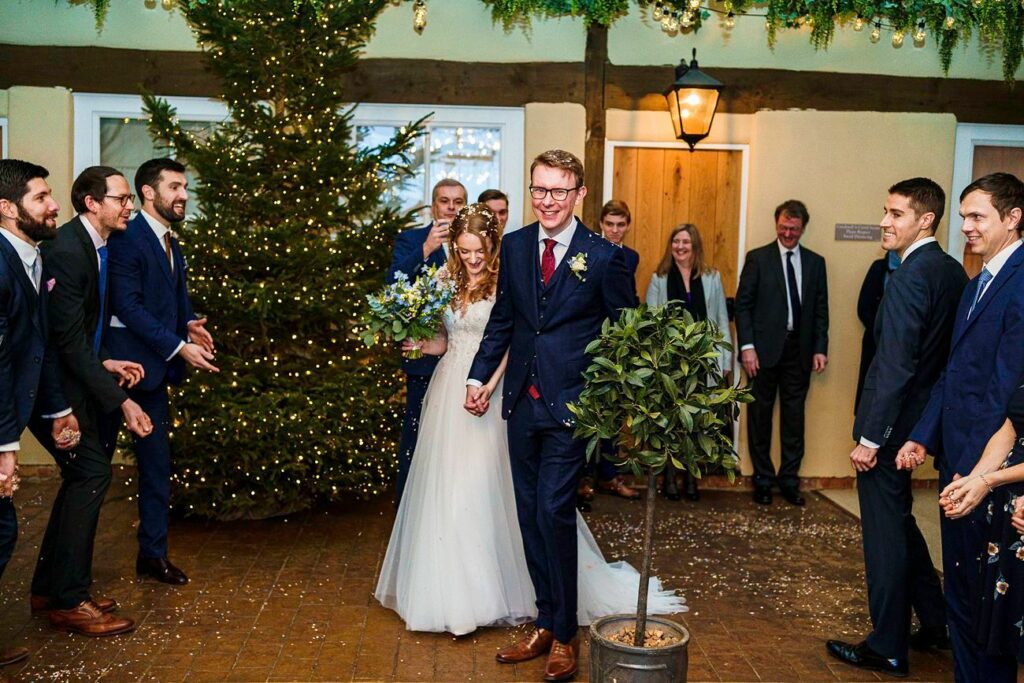 A bride and groom, both smiling, walk hand-in-hand indoors with a decorated Christmas tree nearby. The bride wears a white dress, holding a bouquet, while the groom is in a dark suit. Guests stand on either side, clapping and smiling. Twinkling fairy lights add a festive touch. Image by Documentary Wedding Photographer Lewis Bishop Photography.