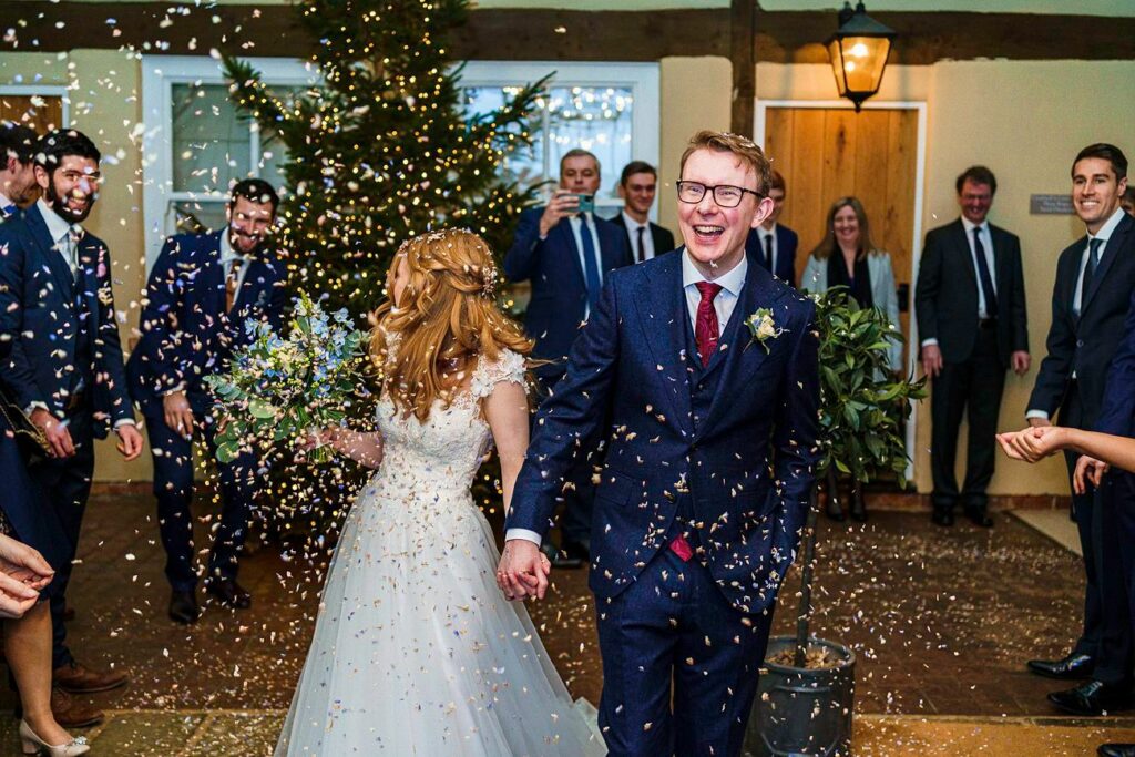 A newlywed couple walks hand-in-hand and smiles as guests throw confetti around them. The bride holds a bouquet and wears a white dress, while the groom is in a navy suit with a red tie. People in the background, dressed formally, cheer and celebrate. Image by Documentary Wedding Photographer Lewis Bishop Photography.