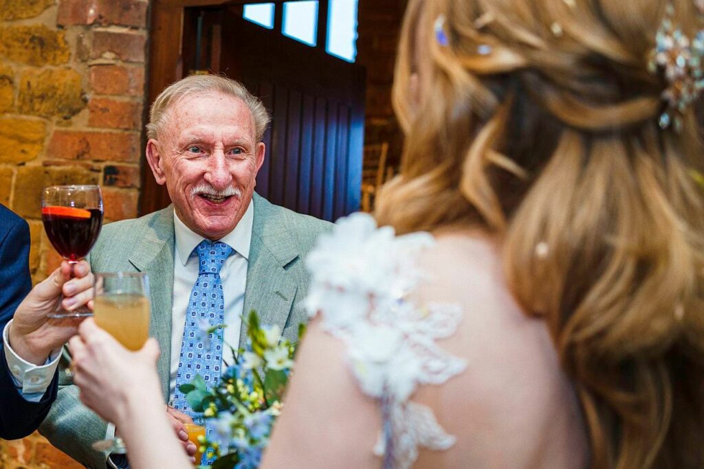 An elderly man in a light grey suit and blue tie, holding a glass of red wine, smiles warmly and clinks glasses with a woman in a white dress with floral detailing. They are indoors, standing near a brick wall and a wooden door. Image by Documentary Wedding Photographer Lewis Bishop Photography.