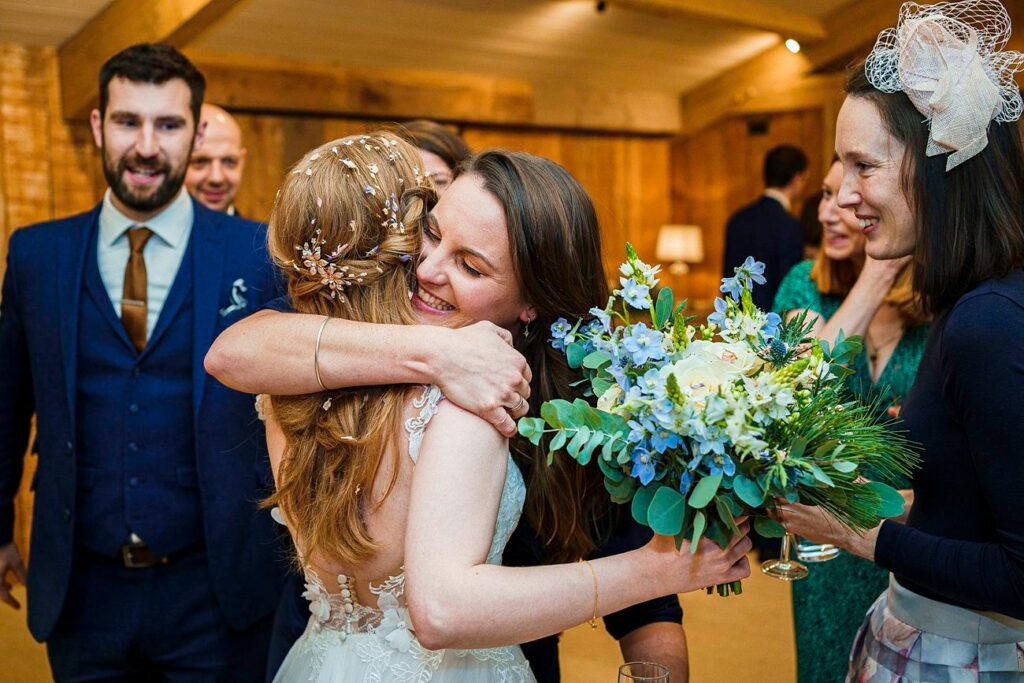A bride with long hair adorned with a floral headpiece hugs a woman holding a bouquet of white and blue flowers. Several people in formal attire stand smiling around them in a warmly lit indoor venue with wooden walls. Image by Documentary Wedding Photographer Lewis Bishop Photography.
