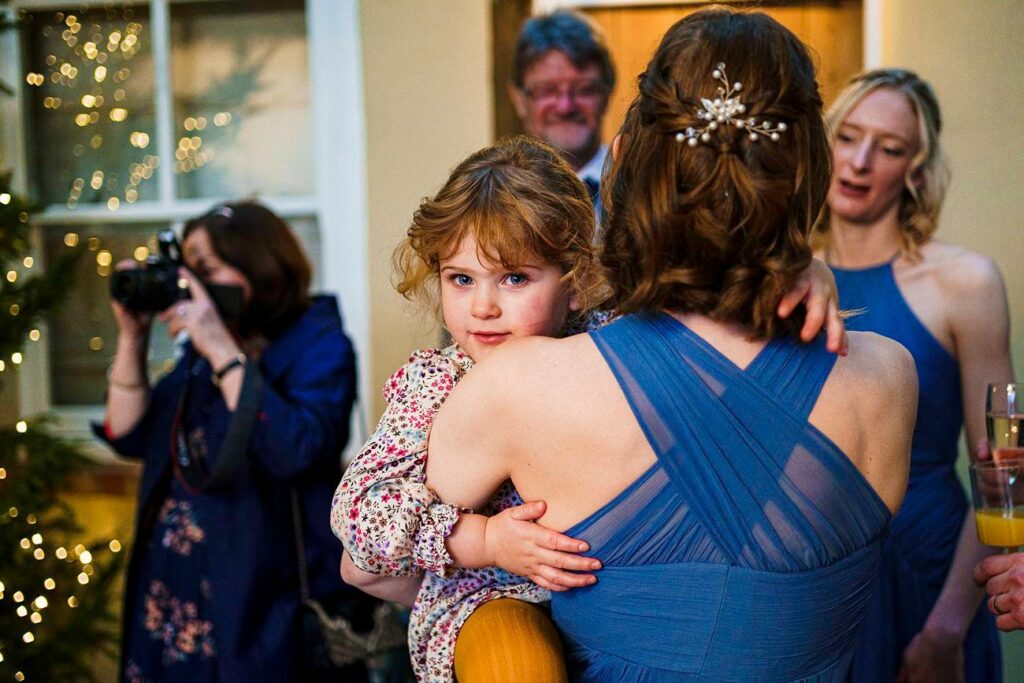 A young girl with curly hair, dressed in a floral print outfit, is being carried by a woman in a blue dress with an intricate back design. They are at an indoor event with warmly lit decor. In the background, a person is taking a photo, and others are engaged in conversation. Image by Documentary Wedding Photographer Lewis Bishop Photography.