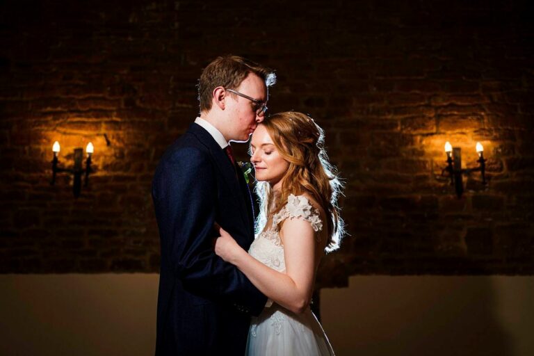 A couple in wedding attire stands close together in a dimly lit room with exposed brick walls and two wall-mounted light fixtures. The groom kisses the bride's forehead while she smiles softly, both illuminated warmly by the ambient lighting. Image by Documentary Wedding Photographer Lewis Bishop Photography.