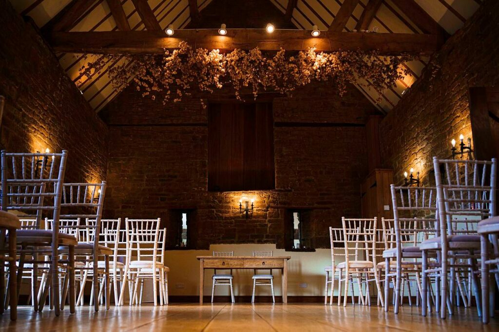 A rustic, well-lit indoor space featuring rows of white wooden chairs arranged for an event. The room has wooden beams on the ceiling, brick walls, and a floral arrangement hanging above. The warm lighting adds to the cozy and inviting ambiance. Image by Documentary Wedding Photographer Lewis Bishop Photography.