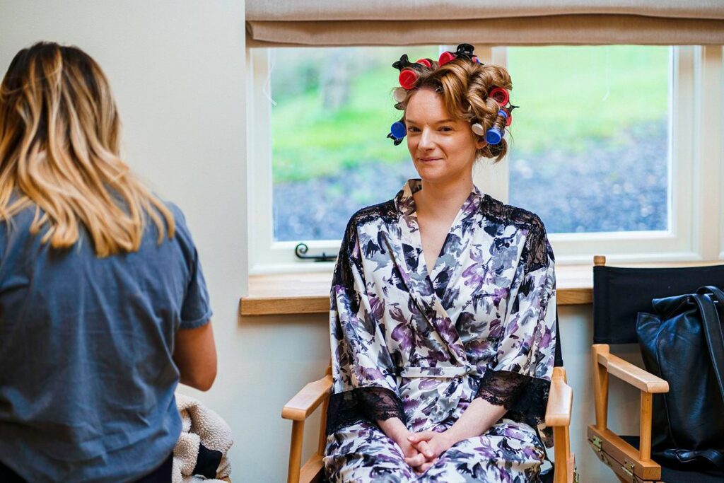 A woman sits on a chair wearing a floral robe with her hair in curlers. She has a slight smile on her face. Another person with long, blonde hair is seen from behind, standing and possibly styling or assisting the seated woman. They are in a room with a window. Image by Documentary Wedding Photographer Lewis Bishop Photography.