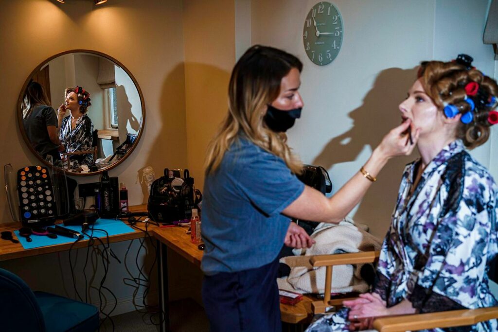 A woman in a patterned robe sits in a chair with hair curlers while a makeup artist, wearing a black face mask, applies makeup. They are positioned in front of a circular mirror on a wooden table with makeup tools, capturing the scene's reflection. Image by Documentary Wedding Photographer Lewis Bishop Photography.