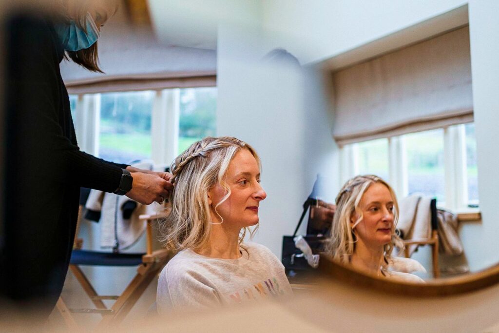 A blonde woman with braided hair sits in front of a mirror while a hairstylist works on her hair. The woman smiles slightly, and the reflection of her happy expression is seen in the mirror. Light floods the room from large windows in the background. Image by Documentary Wedding Photographer Lewis Bishop Photography.