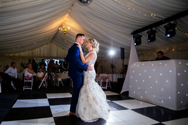 A bride and groom share their first dance in a beautifully decorated wedding tent. The bride wears a flowing white dress, and the groom is in a navy suit. Guests are seated and watching, while a DJ stands at a booth with twinkling lights. Image by Documentary Wedding Photographer Lewis Bishop Photography.