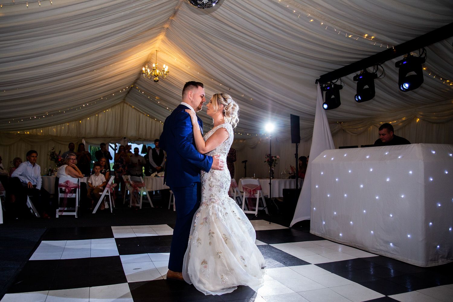 A bride and groom share their first dance in a beautifully decorated wedding tent. The bride wears a flowing white dress, and the groom is in a navy suit. Guests are seated and watching, while a DJ stands at a booth with twinkling lights. Image by Documentary Wedding Photographer Lewis Bishop Photography.