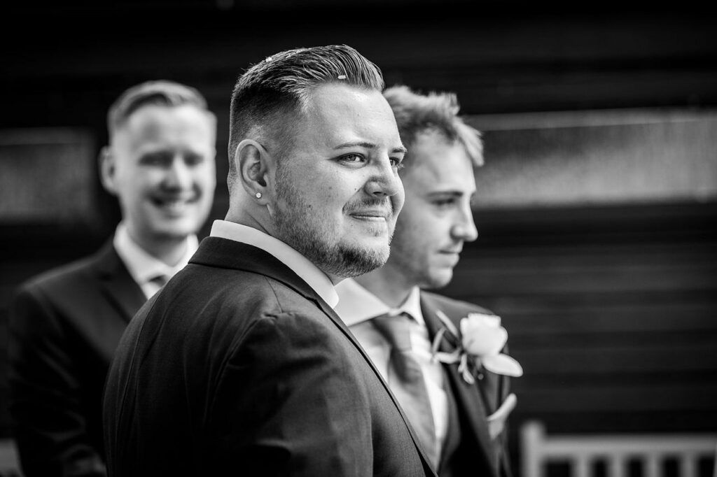 Black and white photo featuring three men in suits at what appears to be a wedding. The man in the foreground has a beard and an earring, and is smiling slightly. The two other men behind him, also dressed formally, are laughing and looking elsewhere. Image by Documentary Wedding Photographer Lewis Bishop Photography.