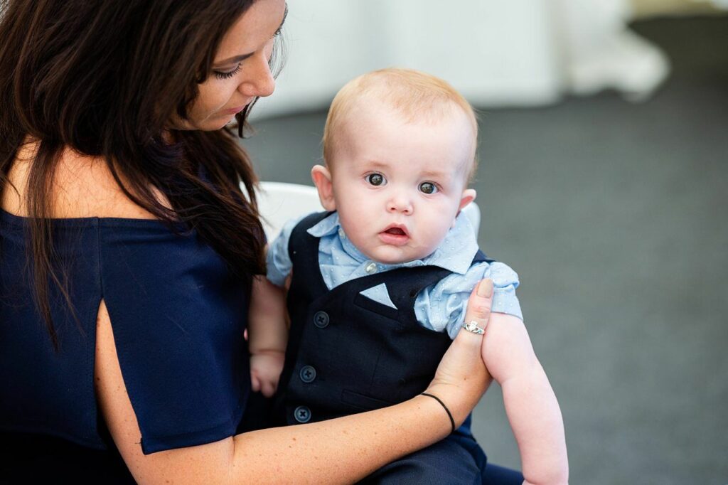 A woman with long brown hair wears a navy blue off-shoulder dress and holds a baby dressed in a blue shirt and black vest. The baby has a curious expression and looks directly at the camera. The background is softly blurred. Image by Documentary Wedding Photographer Lewis Bishop Photography.