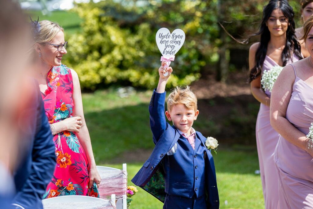 A young boy in a blue suit holds up a heart-shaped sign that reads "Uncle Ryan here comes your bride!" He is walking down an outdoor wedding aisle, surrounded by guests, some of whom are smiling and dressed in colorful attire. Image by Documentary Wedding Photographer Lewis Bishop Photography.