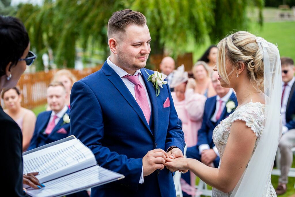 A bride and groom exchange rings during an outdoor wedding ceremony. The groom is in a blue suit with a pink tie, and the bride in a white dress with a veil. An officiant stands to the side holding a book, and guests are seated in the background. Image by Documentary Wedding Photographer Lewis Bishop Photography.