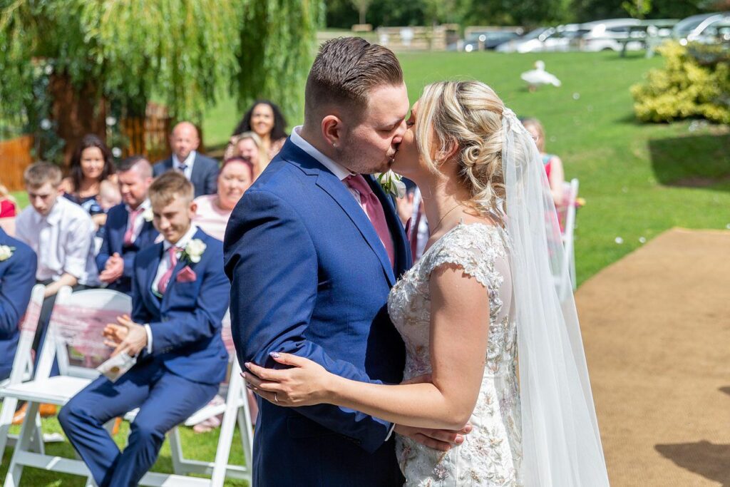 A bride and groom share a kiss in an outdoor wedding ceremony. The bride, in a white lace dress and veil, and the groom, in a blue suit, are surrounded by smiling guests seated on white chairs. Greenery and trees are in the background on a sunny day. Image by Documentary Wedding Photographer Lewis Bishop Photography.