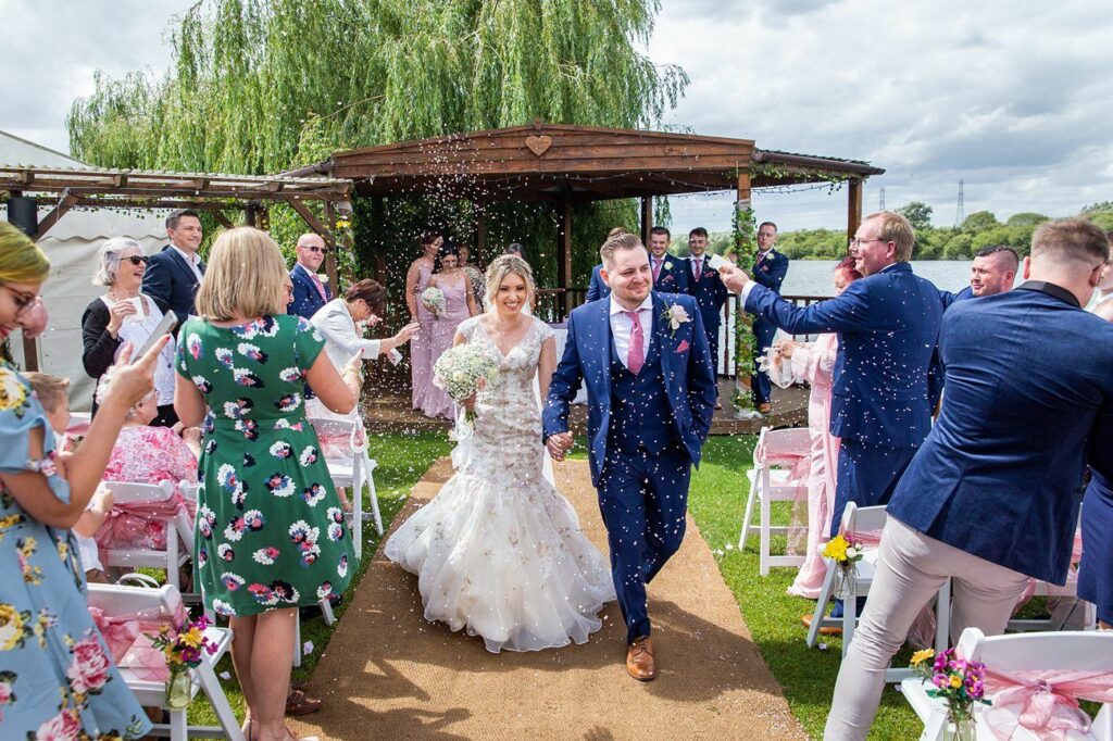A newly married couple walks down the aisle outdoors, smiling as guests celebrate by throwing confetti. The bride wears a white gown and holds a bouquet, while the groom is in a blue suit with a pink tie. The guests are seated and standing, applauding under a wooden pergola. Image by Documentary Wedding Photographer Lewis Bishop Photography.
