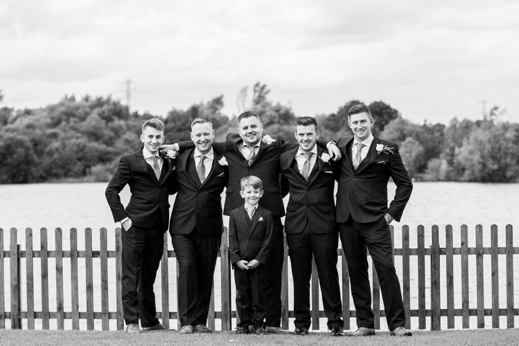 A black-and-white photo shows five men in suits standing with their arms around each other, smiling in front of a wooden fence and a lake. A young boy in a suit stands in front of them, also smiling. Trees and a cloudy sky are visible in the background. Image by Documentary Wedding Photographer Lewis Bishop Photography.