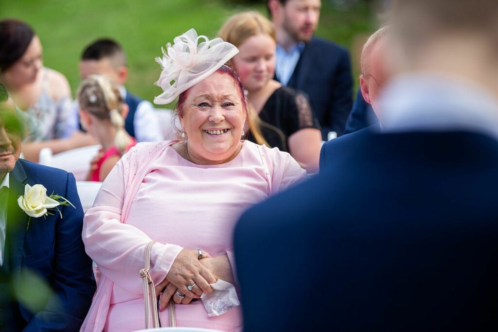 A woman in a pink outfit and matching hat, holding a purse, smiles while seated outdoors at an event. People are seated around her, and a person in a navy suit is in the foreground, out of focus. The background shows a green, grassy area. Image by Documentary Wedding Photographer Lewis Bishop Photography.