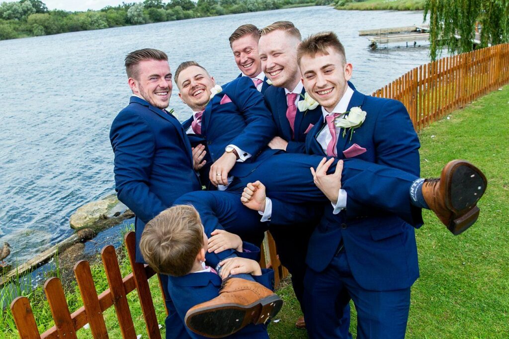 A group of men dressed in matching blue suits with pink ties and white flower boutonnieres are laughing together near a lakeside. They are lifting one of their friends in the air playfully. The scene is joyful and the background features a wooden fence and water. Image by Documentary Wedding Photographer Lewis Bishop Photography.