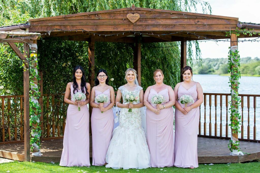 A bride in a white gown stands under a wooden gazebo with four bridesmaids in matching light pink dresses. They are all holding small bouquets, and there is a lake and greenery in the background. The scene is bright and cheerful, suggesting a wedding ceremony. Image by Documentary Wedding Photographer Lewis Bishop Photography.