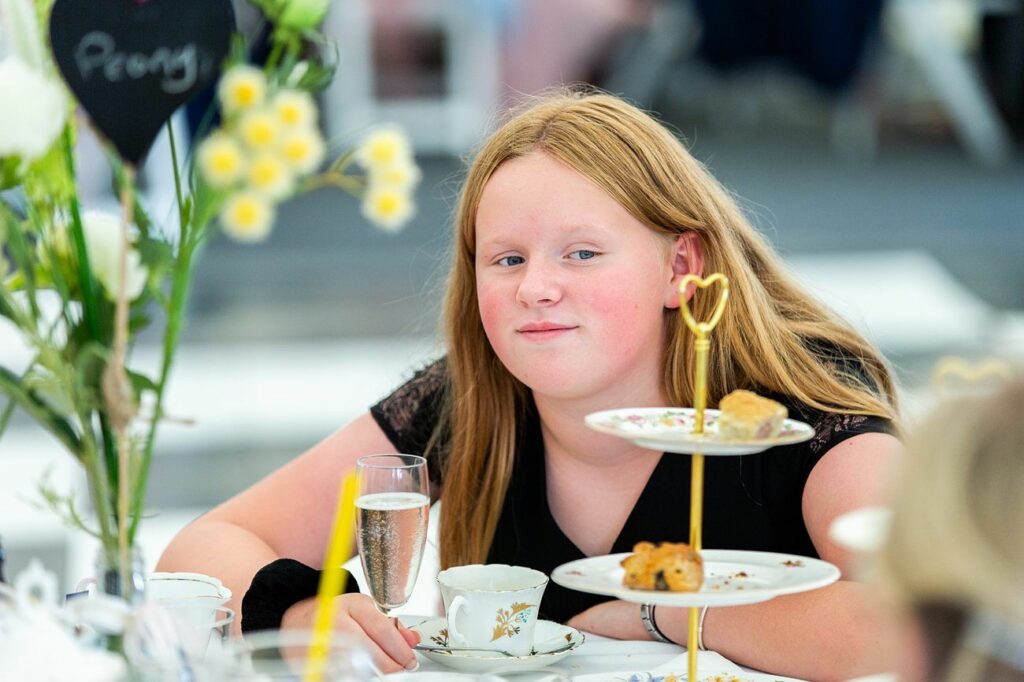 A young person with long red hair is seated at a table, gazing ahead with a relaxed expression. In front of them, there's a tiered tray with scones and pastries, a tea cup, and a glass of sparkling beverage. The table has floral decorations and a heart-shaped sign. Image by Documentary Wedding Photographer Lewis Bishop Photography.