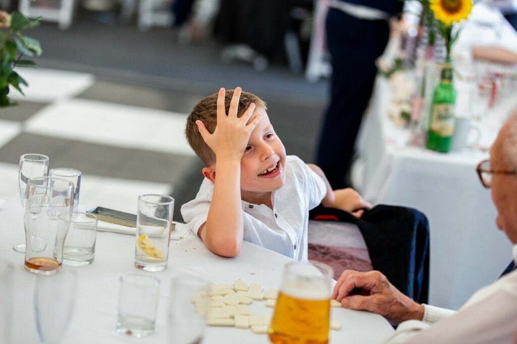 A young boy with an amused expression sits at a table with scattered puzzle pieces, placing his hand on his forehead as if reacting to something funny. An elderly man is seated across from the boy, and the table has a beer glass and empty drink glasses. Image by Documentary Wedding Photographer Lewis Bishop Photography.