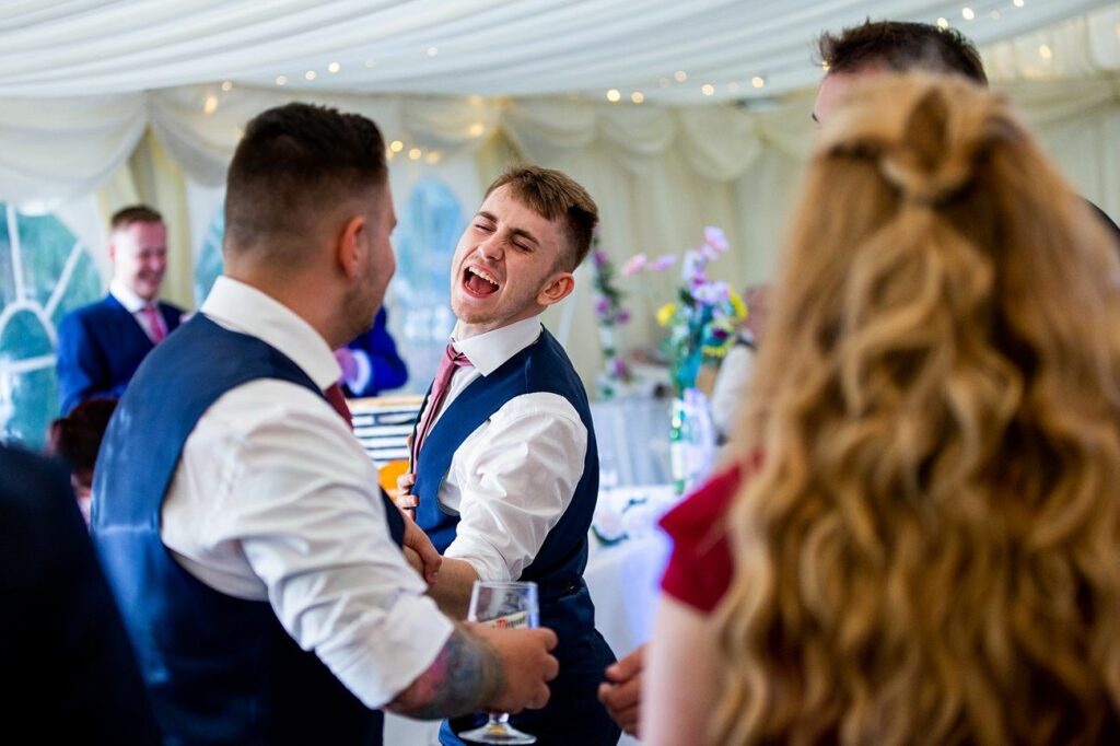 A group of people, dressed formally, are enjoying themselves at an event. One man, laughing joyfully, is in the center with a glass of drink and engaged in conversation. The tent is decorated with pleated drapes and fairy lights, and there are floral arrangements in the background. Image by Documentary Wedding Photographer Lewis Bishop Photography.