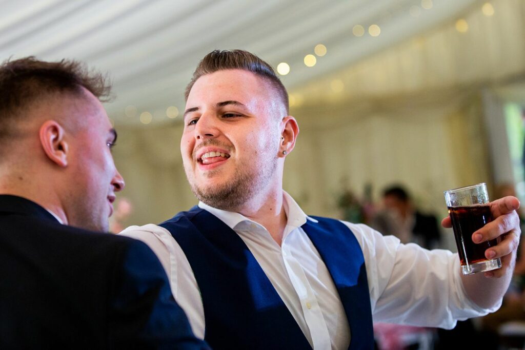 A man wearing a navy vest and white shirt is joyfully holding a drink, engaged in conversation with another man in a dark suit. They appear to be at an indoor celebration with festive lighting in the background. Image by Documentary Wedding Photographer Lewis Bishop Photography.
