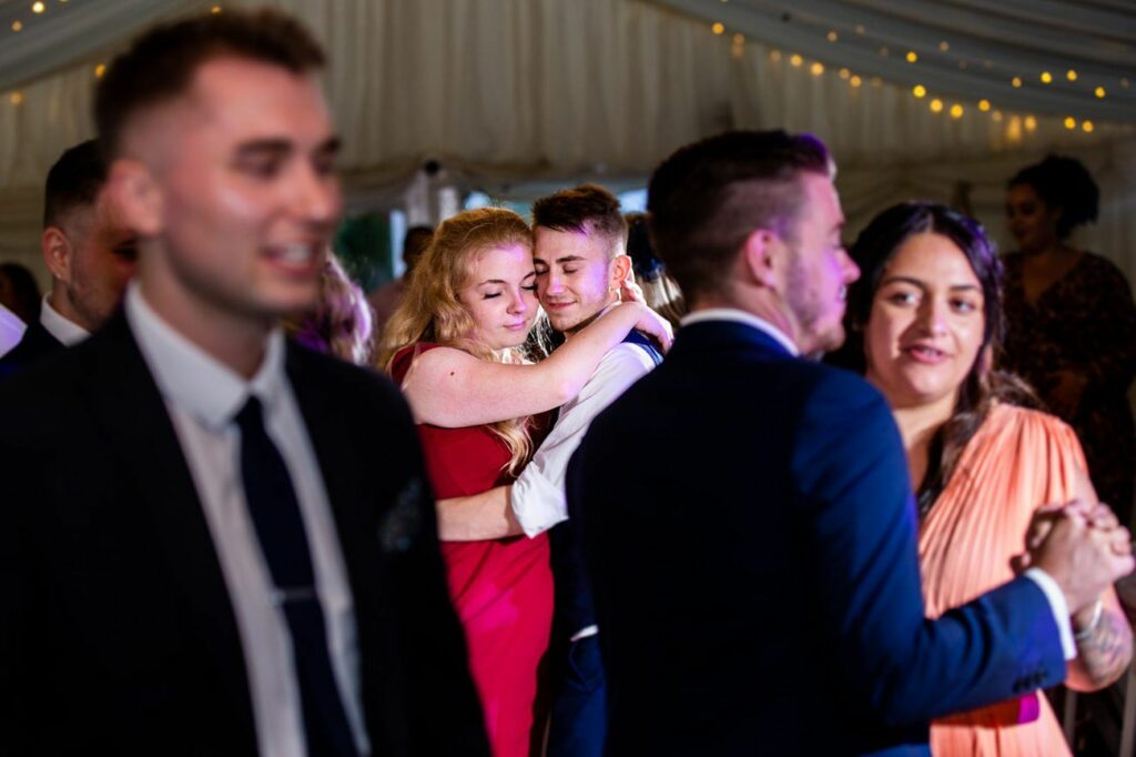 People are dancing at an event in a tent with string lights. A couple in the center embraces, enjoying the moment. Other couples dance nearby, with soft lighting adding to the festive atmosphere. One person in the foreground is slightly out of focus. Image by Documentary Wedding Photographer Lewis Bishop Photography.