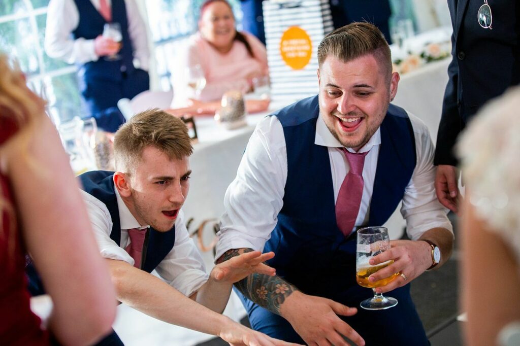Two men in vests and ties are having fun and dancing at a lively event, possibly a wedding reception. One is crouching while laughing and holding a drink, and the other is gesturing animatedly. People in semi-formal attire and decorations are visible in the background. Image by Documentary Wedding Photographer Lewis Bishop Photography.