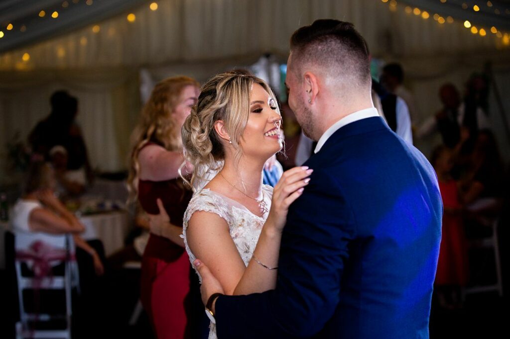 A couple wearing formal attire shares a dance in a warmly lit room. The woman, in a white dress, smiles and gazes at her partner, who is in a dark suit. Guests in the background watch, some seated at tables decorated with lights. Image by Documentary Wedding Photographer Lewis Bishop Photography.