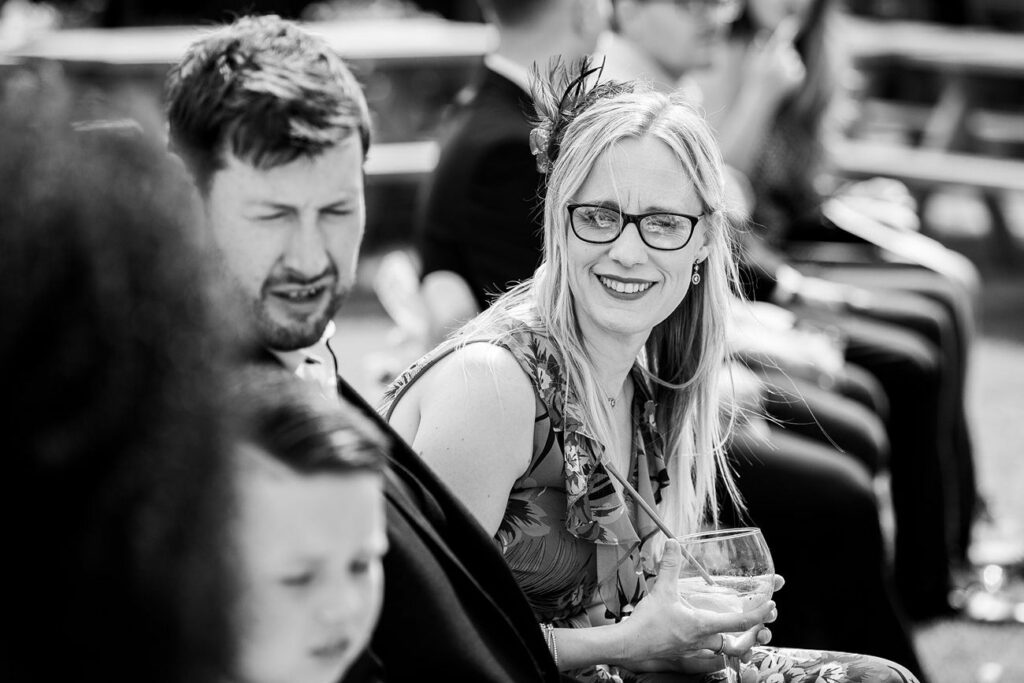 A black and white image shows a group of people seated outdoors. A woman with glasses and a floral dress smiles while holding a drink. A man next to her looks forward, and a young child is partially visible in the foreground. The background is slightly blurred. Image by Documentary Wedding Photographer Lewis Bishop Photography.