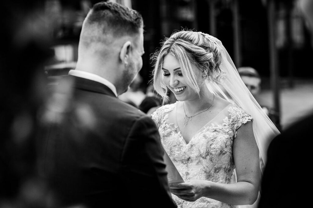 A black-and-white photo of a bride and groom facing each other during their wedding ceremony. The bride, wearing a lace dress and veil, smiles as she looks at the groom. The groom is in a suit, seen from the back. Guests are blurred in the background. Image by Documentary Wedding Photographer Lewis Bishop Photography.