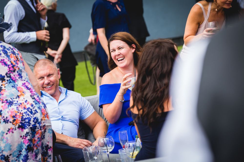 A group of people socializing at an outdoor event. A woman in a blue dress is laughing with a drink in hand, sitting next to a man in a light blue shirt. Other guests are seen mingling in the background. The scene appears lively and joyous. Image by Documentary Wedding Photographer Lewis Bishop Photography.