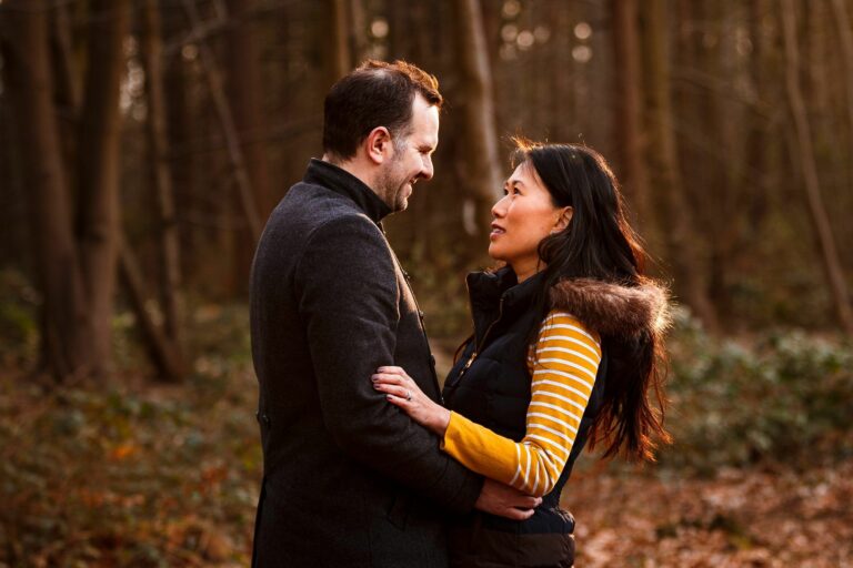 A couple gazes into each other's eyes while standing among trees in a forest. They are both dressed warmly; the man wears a coat, and the woman wears a striped shirt under a vest with fur trim. The ground is covered in fallen leaves, and the sunlight filters through the trees. Image by Documentary Wedding Photographer Lewis Bishop Photography.