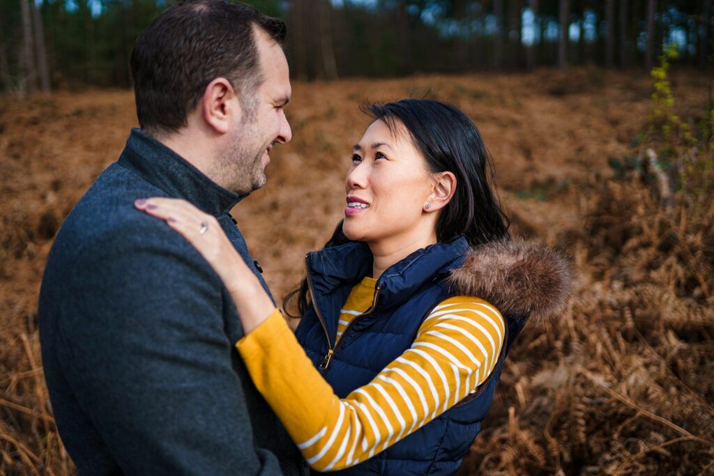 A couple stands outside in a wooded area, smiling warmly at each other. The woman, wearing a yellow and white striped shirt with a blue vest, has her hand on the man’s shoulder. The man, wearing a gray coat, looks back at her affectionately. Image by Documentary Wedding Photographer Lewis Bishop Photography.
