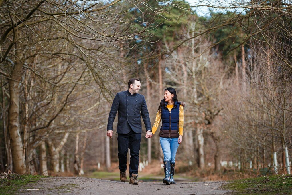 A couple holding hands and smiling at each other while walking on a wooded path. The man is wearing a dark coat and trousers, and the woman is wearing a yellow sweater, a vest, and jeans with boots. Bare trees line the path, and tall trees stand in the background. Image by Documentary Wedding Photographer Lewis Bishop Photography.