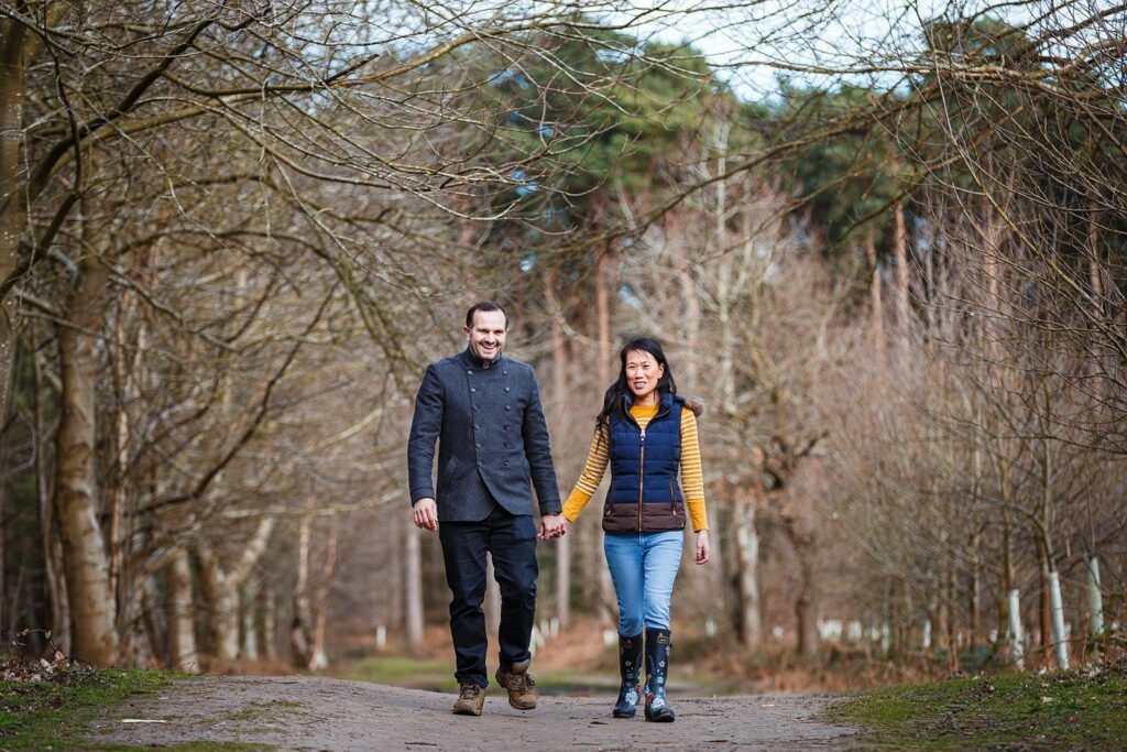 A couple is walking, holding hands, on a dirt path through a forest with leafless trees. The man is dressed in a dark jacket and trousers, while the woman wears a yellow sweater, blue jeans, and a dark vest. Both appear happy, surrounded by a peaceful, natural setting. Image by Documentary Wedding Photographer Lewis Bishop Photography.