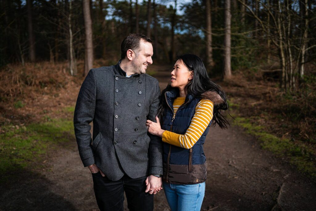 A couple stands on a forest path, looking at each other and smiling. The man, with short hair, wears a gray coat and black pants, while the woman, with long dark hair, wears a striped yellow shirt and a blue vest. Trees and foliage surround them. Image by Documentary Wedding Photographer Lewis Bishop Photography.