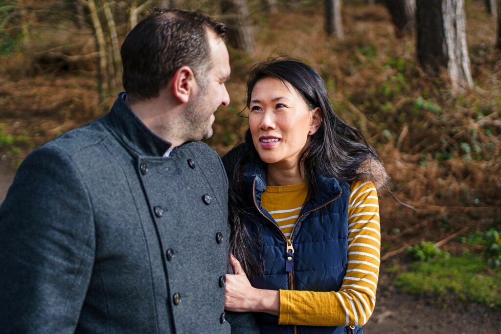 A couple walks through a wooded area, gazing at each other. The man wears a gray coat, and the woman, in a yellow and white striped shirt and navy vest, holds his arm. The background is lined with trees and brown foliage. Image by Documentary Wedding Photographer Lewis Bishop Photography.