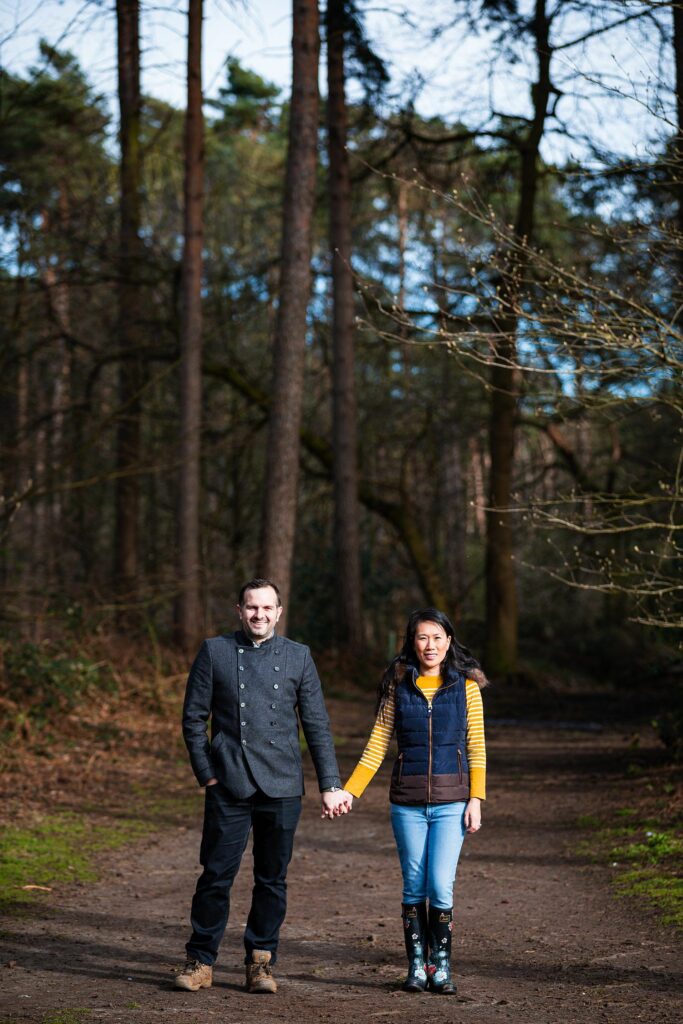 A man and woman hold hands while walking on a forest path. The man is wearing a dark coat and black pants, and the woman is in a yellow and black jacket, jeans, and floral-patterned rain boots. Tall trees and foliage surround them in the serene setting. Image by Documentary Wedding Photographer Lewis Bishop Photography.