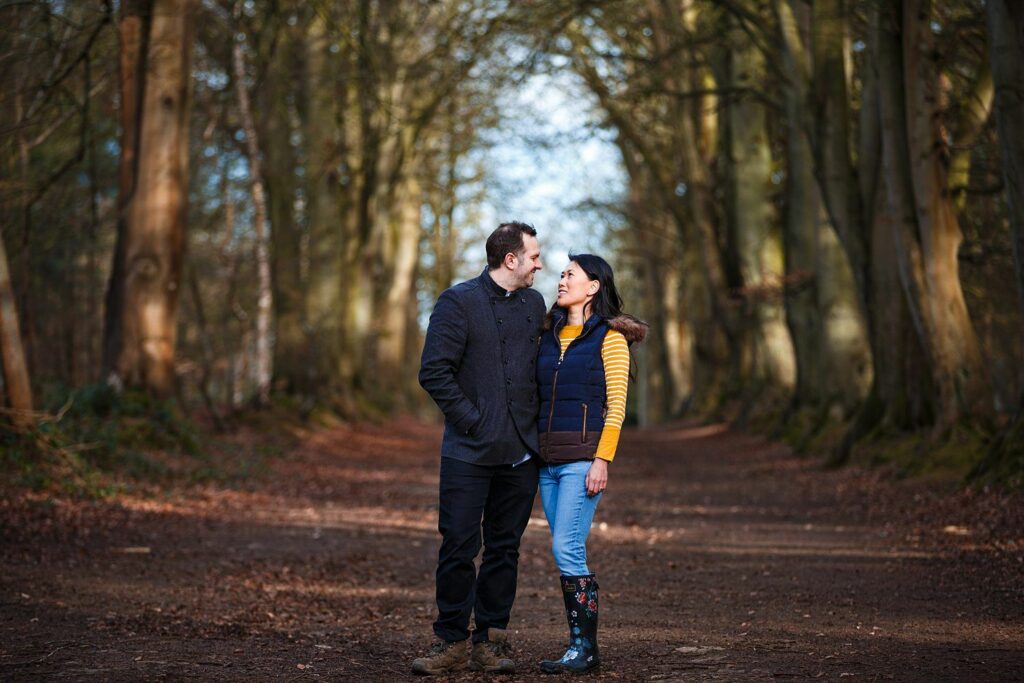 A couple standing in a forest path, facing each other and holding hands. The man wears a dark coat and black pants, while the woman is dressed in a blue vest, yellow sweater, jeans, and knee-high boots. Sunlight filters through the trees overhead. Image by Documentary Wedding Photographer Lewis Bishop Photography.