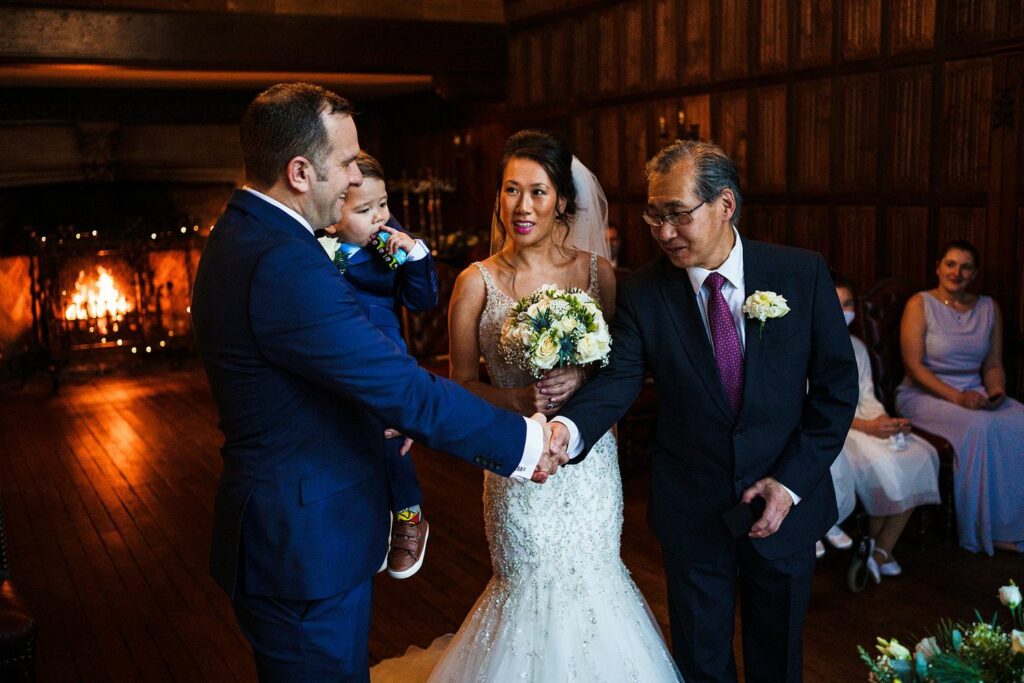 A groom shakes hands with an older man as he holds a toddler boy, while a bride in a white wedding dress stands next to them holding a bouquet. Several seated guests can be seen in the background, and a fireplace adds warmth to the wooden interior room. Image by Documentary Wedding Photographer Lewis Bishop Photography.