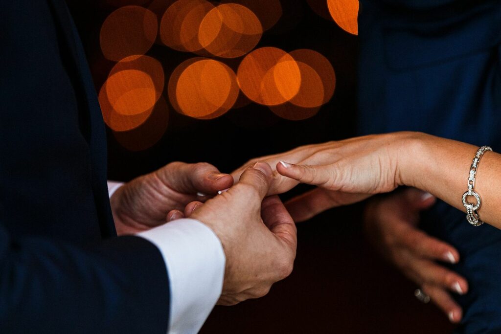 Close-up of a person in a dark suit placing a ring on another person's finger, against a backdrop of blurred orange and yellow lights. The person receiving the ring is wearing a bracelet. Image by Documentary Wedding Photographer Lewis Bishop Photography.