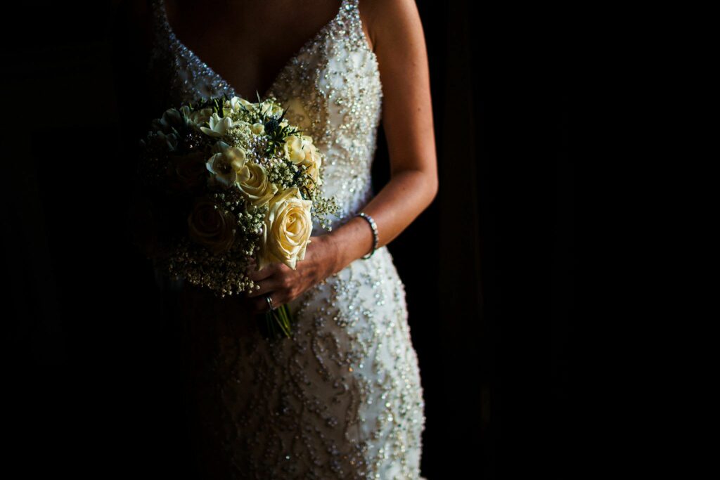 A bride in a beaded, white wedding gown holds a bouquet of white and pale yellow roses with baby's breath, standing against a dark backdrop. The focus is on the flowers and the intricate details of the dress, while her face remains unseen. Image by Documentary Wedding Photographer Lewis Bishop Photography.