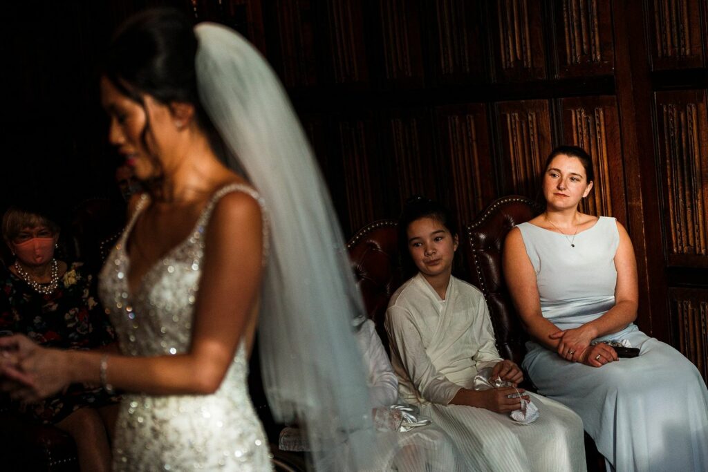 A bride in a white beaded gown and veil stands in the foreground. In the background, three people are seated, including a young girl in white and an adult woman in a light blue dress, along with another person partially visible. The setting features dark wooden paneling. Image by Documentary Wedding Photographer Lewis Bishop Photography.
